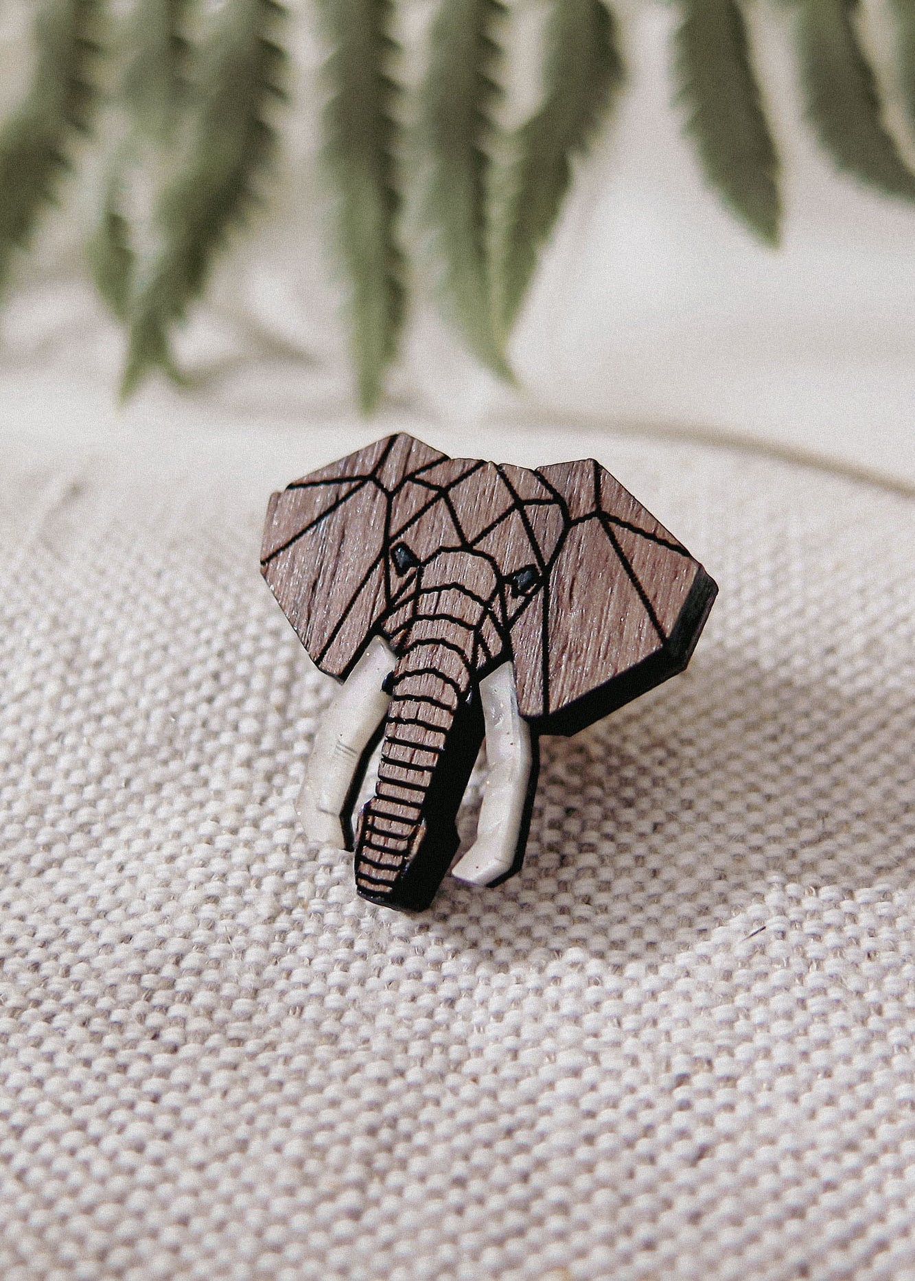 A handcrafted Elephant Pin with a geometric design and white tusks rests on textured beige fabric. Softly blurred green fern leaves appear in the background.