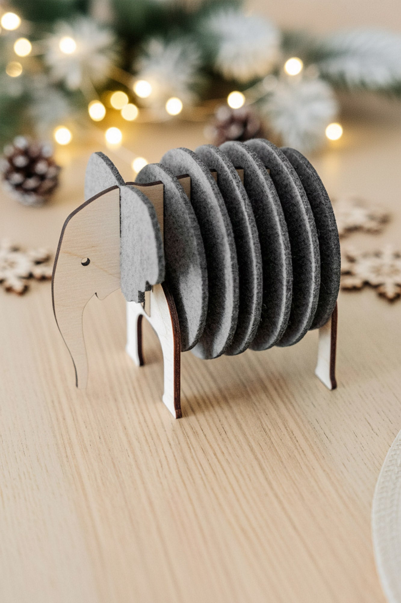 A wooden and felt elephant coasters holder sits on a light table, adding witty design and furniture protection, with pinecones, snowflake decorations, and string lights blurred in the background.