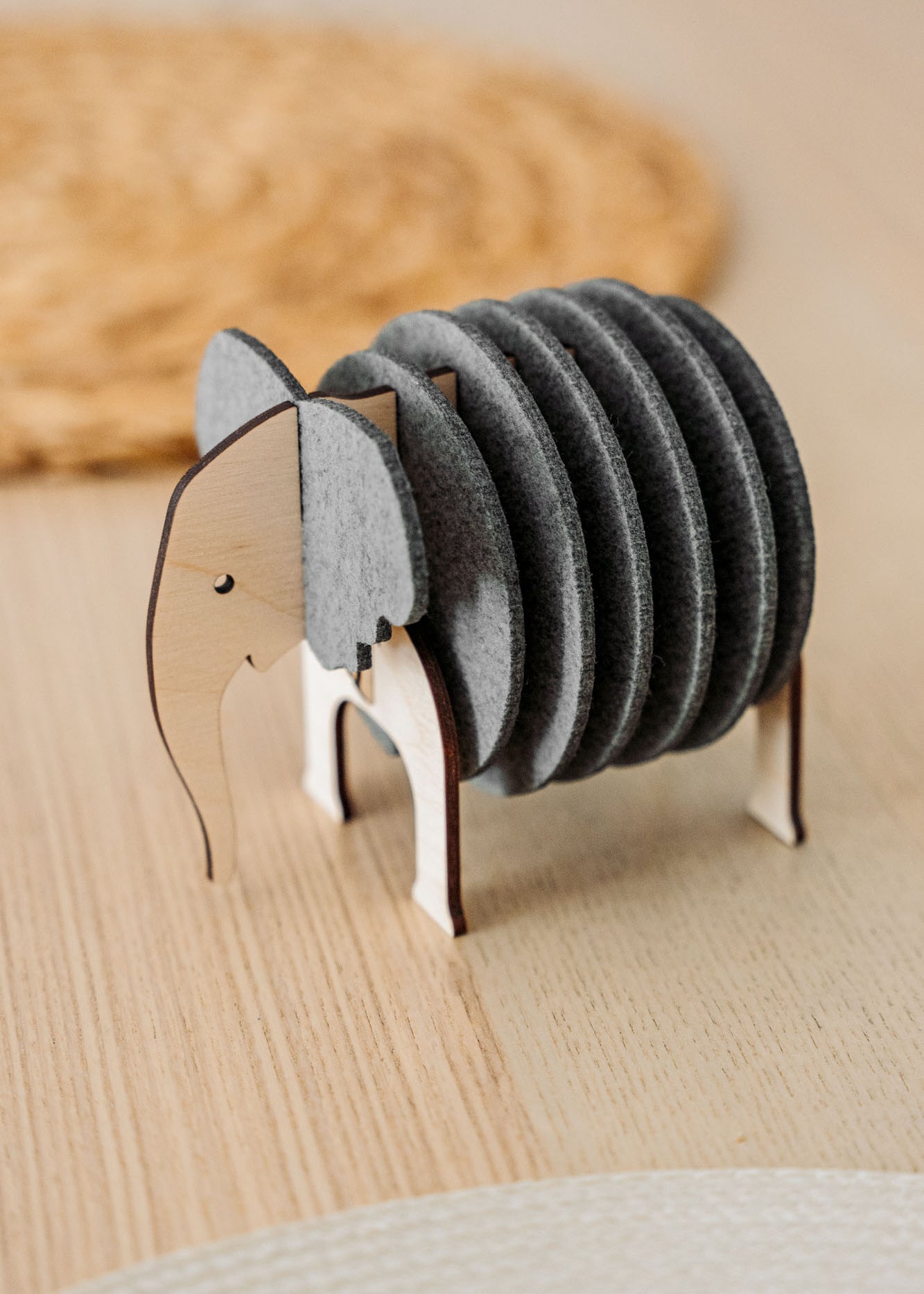 A wooden and felt Elephant Coasters holder with witty design stands on a wooden table, offering both charm and furniture protection, while a round woven placemat is blurred in the background.
