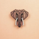A handcrafted Elephant Pin showcases a geometric wooden carving of an elephants head with white tusks, centered on a light peach background.