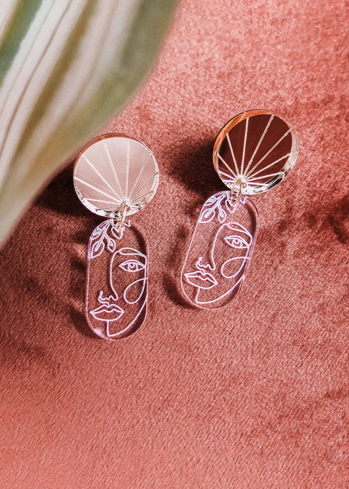 Silver handcrafted Face Earrings with abstract designs and round, reflective tops rest on a soft pink surface. A green and white leaf partially frames the image, adding artful sophistication to elevate your style.