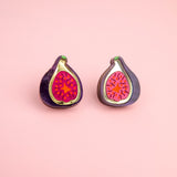Two enamel pins shaped like halved figs with bright pink and red centers rest on a light pink background, perfect for fans of fig jewelry or those who love unique laser-cut studs.