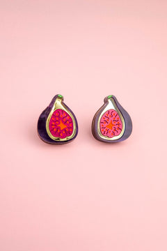 Two enamel pins shaped like halved figs with bright pink and red centers rest on a light pink background, perfect for fans of fig jewelry or those who love unique laser-cut studs.