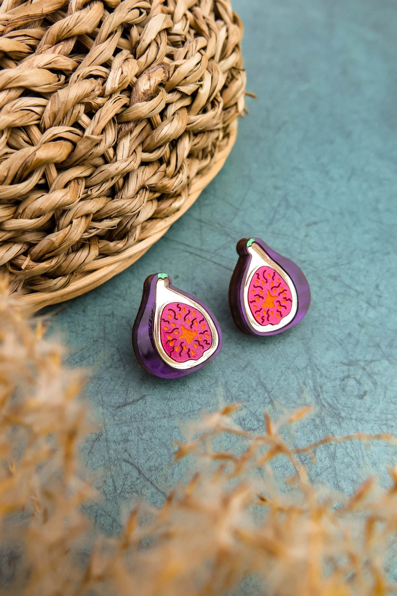 A pair of fig-shaped, hand-painted earrings with purple and pink detailing are displayed on a green surface next to a woven basket. Blurred beige plant stems are visible in the foreground.