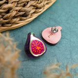 A pair of fig-shaped, hand-painted earrings—one with a detailed purple and pink fig design, the other revealing a plain wooden back and metal post—rest on a textured teal surface beside a woven basket, perfect for lovers of unique fig jewelry.