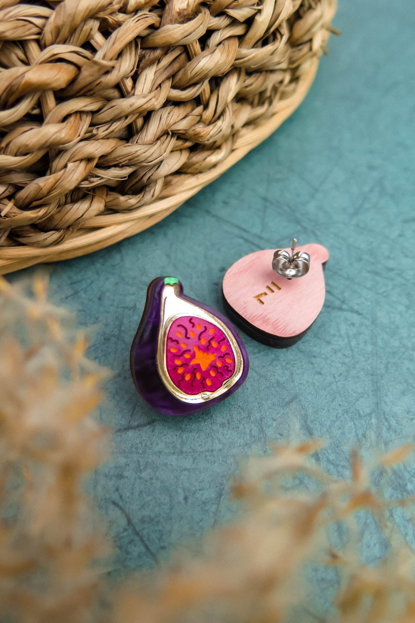 A pair of fig-shaped, hand-painted earrings—one with a detailed purple and pink fig design, the other revealing a plain wooden back and metal post—rest on a textured teal surface beside a woven basket, perfect for lovers of unique fig jewelry.