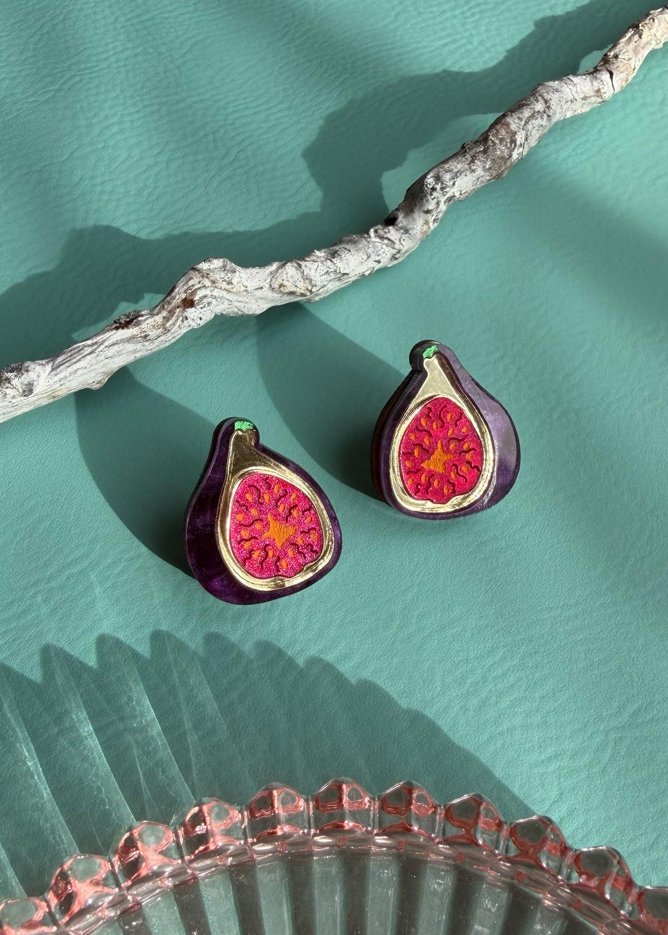 A pair of deep purple earrings, hand-painted to resemble figs with vibrant red centers, rest on a textured teal surface beside a white branch and a pink glass dish.
