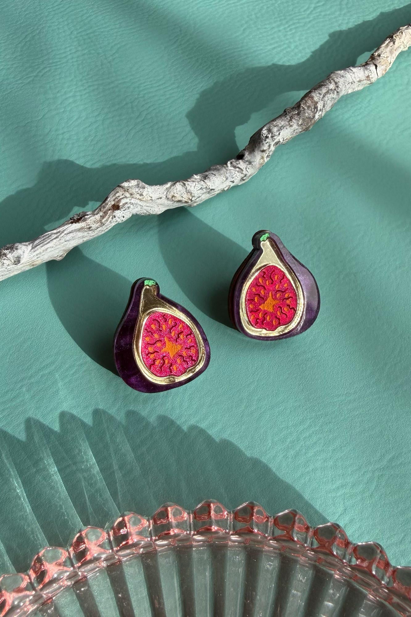 A pair of deep purple earrings, hand-painted to resemble figs with vibrant red centers, rest on a textured teal surface beside a white branch and a pink glass dish.
