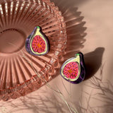 Two fig-shaped laser-cut studs with vibrant pink and deep purple detailing rest on a pink glass dish, casting shadows on a pale pink surface. Some delicate, dried grass lies nearby, adding texture to the scene.