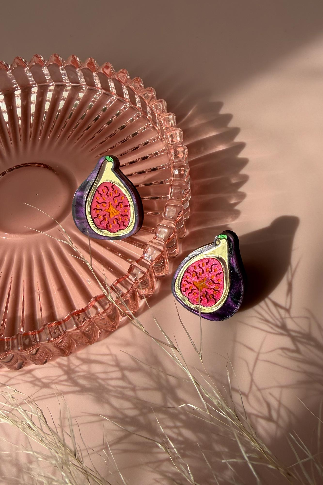 Two fig-shaped laser-cut studs with vibrant pink and deep purple detailing rest on a pink glass dish, casting shadows on a pale pink surface. Some delicate, dried grass lies nearby, adding texture to the scene.