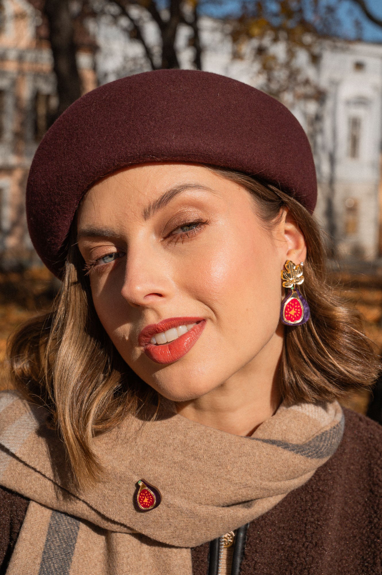 A woman wearing a burgundy beret and a beige scarf poses outdoors. She wears red lipstick, handcrafted fig earrings, and a matching pin. Autumn trees and a building create a scenic background.