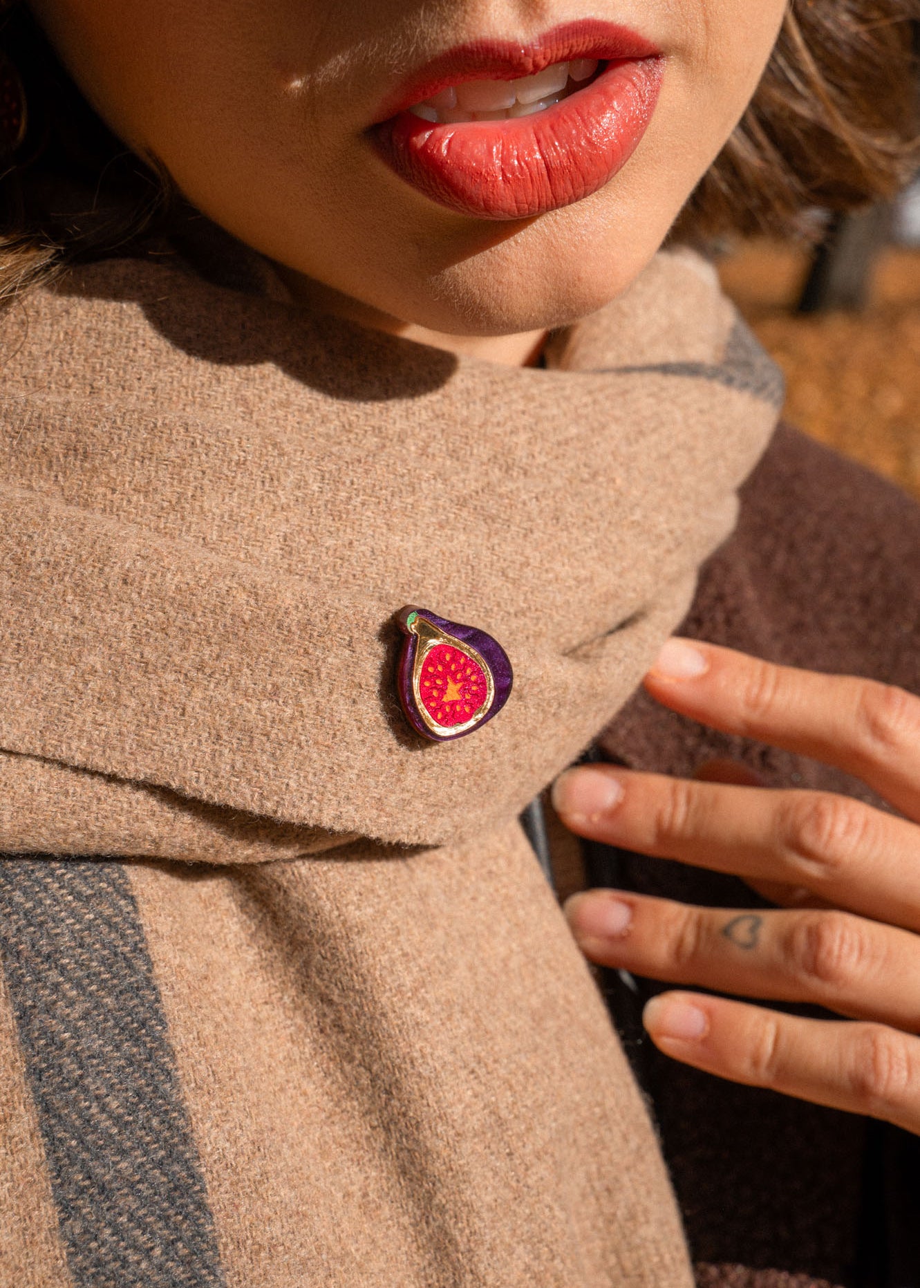 A person wearing red lipstick and a beige scarf with a handcrafted Fig Pin, a unique accessory shaped like a fig; their hand rests on the scarf, showing neat nails and a small finger tattoo.