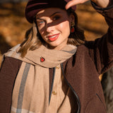 A woman in a brown beret and coat smiles at the camera, holding one hand up to shield her eyes from the sunlight. She wears a beige scarf, red earrings, and a unique accessory—a handcrafted Fig Pin—with autumn leaves blurred in the background.