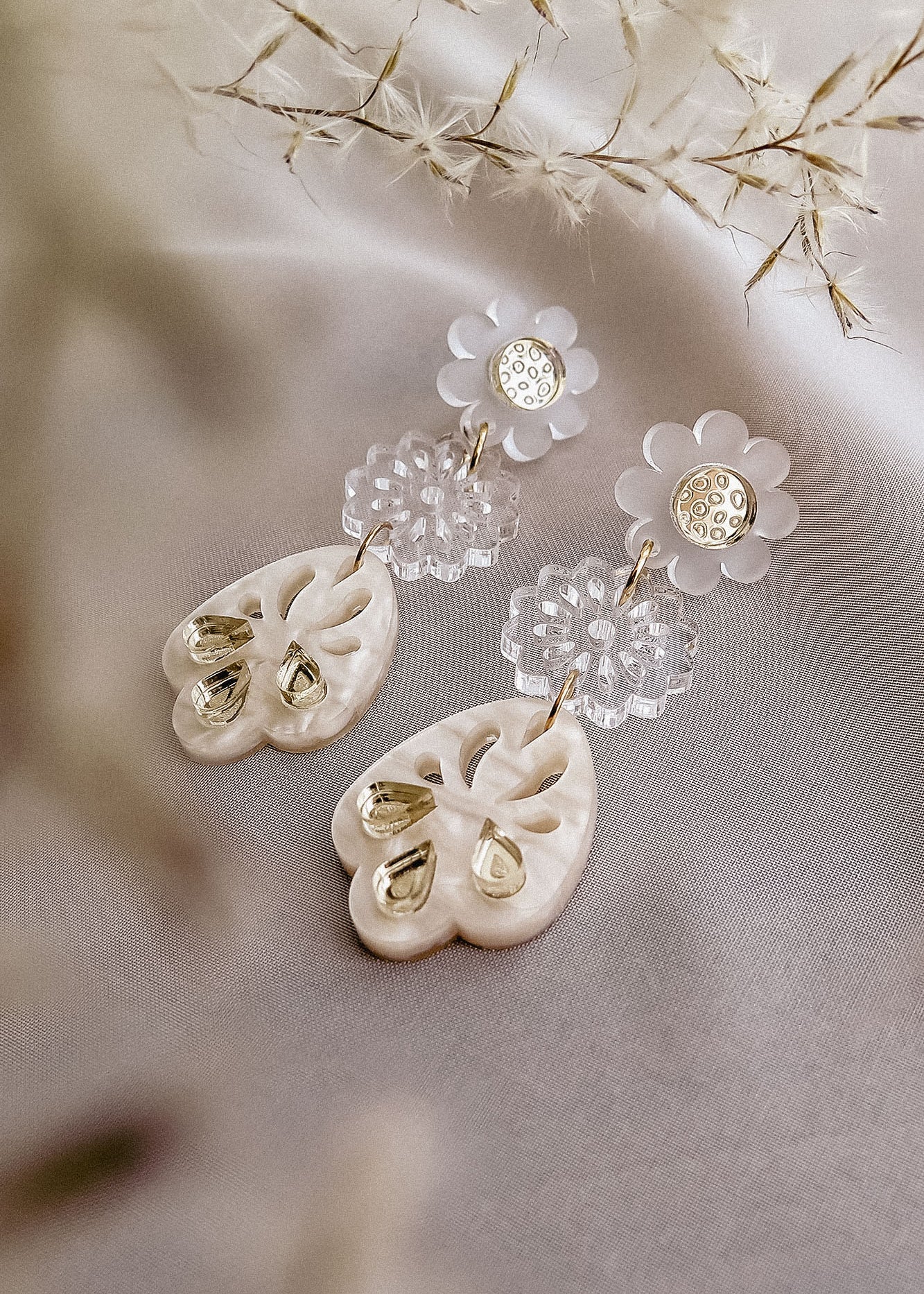 A pair of elegant, handcrafted boho earrings featuring flower-shaped clear studs and ornate, heart-shaped white pendants with gold accents, displayed on soft fabric with delicate dried flowers nearby.