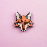 A geometric Fox Pin brooch, crafted from recycled wood in orange, black, and white hues, is displayed on a pink background—perfect for an eco-conscious style.