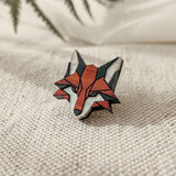 A geometric Fox Pin crafted from recycled wood in red, black, and white rests on textured cream fabric, with blurred green fern leaves adding an eco-conscious style to the scene.