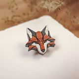 A geometric Fox Pin brooch, crafted from recycled wood in orange, black, and white tones, rests on a white surface with blurred dried plants in the background—a perfect touch for an eco-conscious style.