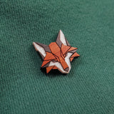 A geometric Fox Pin crafted from recycled wood, featuring orange, white, and brown details, is attached to a green fabric background—perfect for showcasing your eco-conscious style.