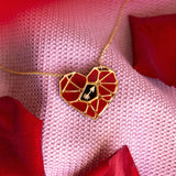 A handcrafted gold heart pendant, featuring a red mosaic design and a small white hand symbol, rests on pink textured fabric amid red rose petals—a striking Fragment of Love Necklace.