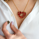 A person wearing a white shirt displays the handcrafted Fragment of Love Necklace featuring a red, geometric heart pendant and holds a small black pin with a gold arrow design. Only the lower face and hand are visible.