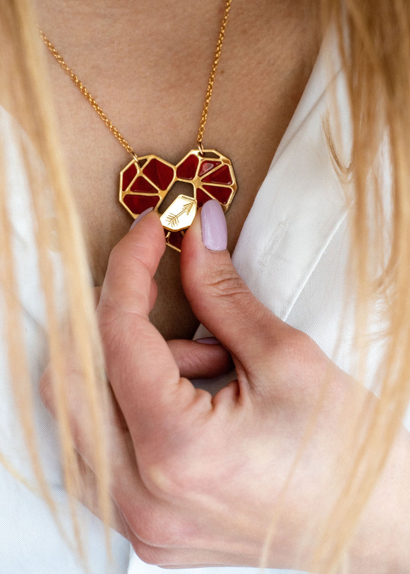 A person with light pink nail polish holds a handcrafted gold and red heart pendant, the Fragment of Love Necklace, against their chest, visible above a white shirt.