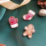 Three decorated holiday cookies—a mitten, a stocking, and a gingerbread Man Pin—are arranged on green fabric with gold ribbon, pinecones, and glittery ornaments, adding a festive touch to your Christmas party display.