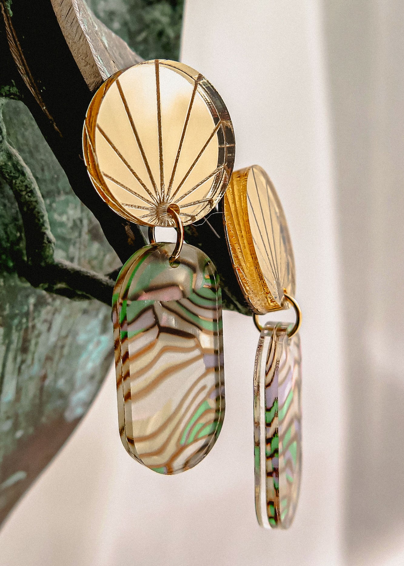 Close-up of handcrafted gold earrings with engraved fan-patterned tops and elongated oval dangles made of translucent abalone, showcasing wavy green, brown, and cream stripes.