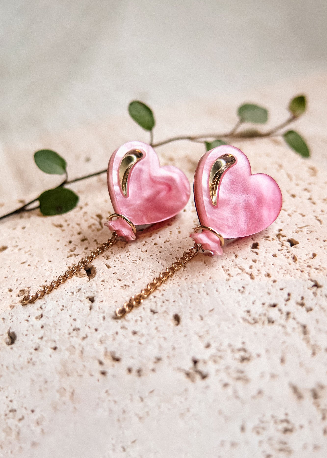 A pair of pink Heart Balloon Studs with gold accents and dangling chains, displayed on a textured cream surface with a green leafy stem in the background—perfect for adding vibrant style to any look.