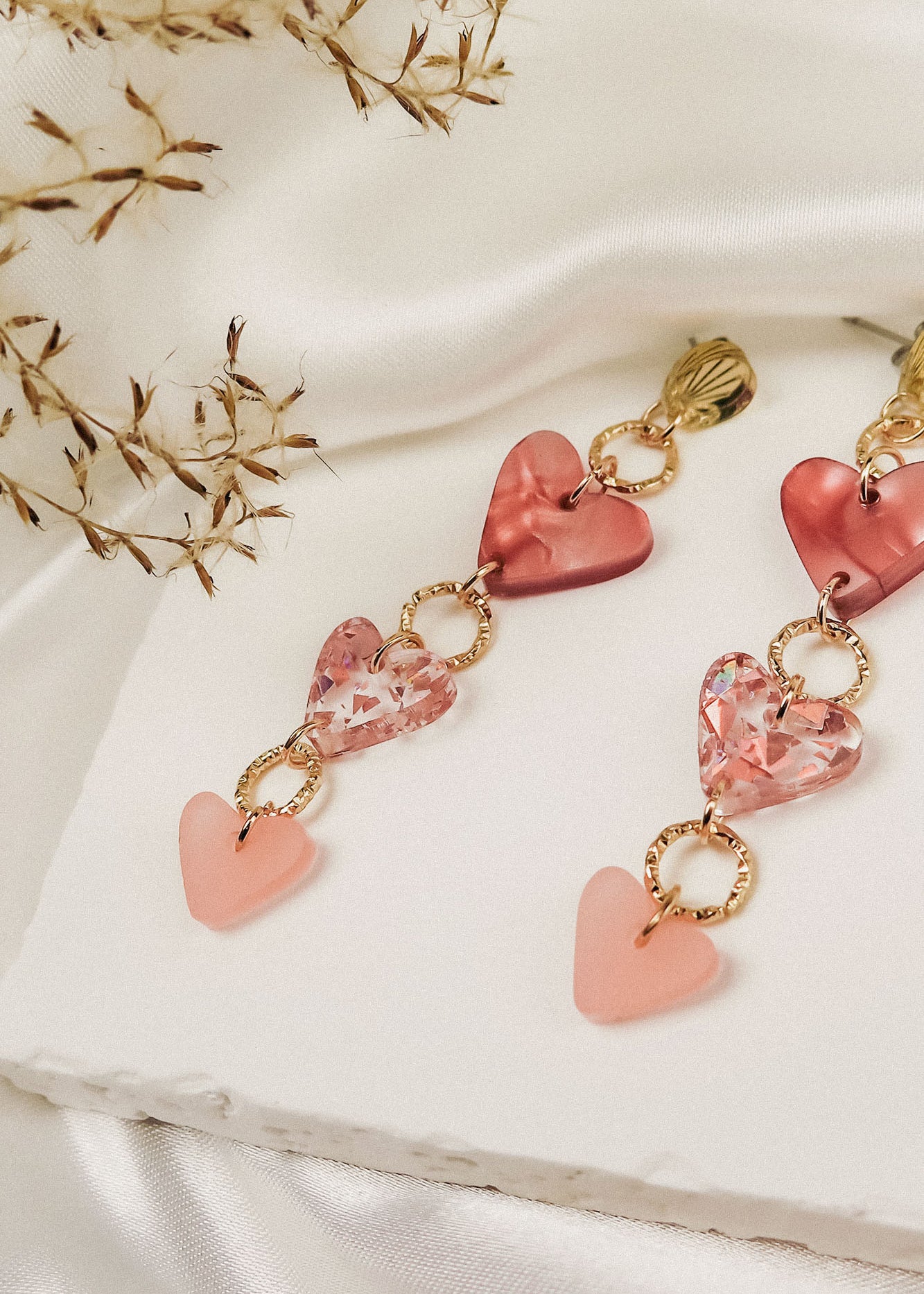 A pair of Heart Chain Earrings with gold links and pink heart-shaped charms, displayed on a white textured surface with delicate dried flowers and draped ivory fabric in the background, adds a touch of allergy-free comfort to your look.