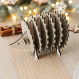 A wooden hedgehog figurine, perfect for home decor, sits on a light wooden table. In the blurred background, lights, fir branches, and cinnamon sticks create a festive, cozy atmosphere.