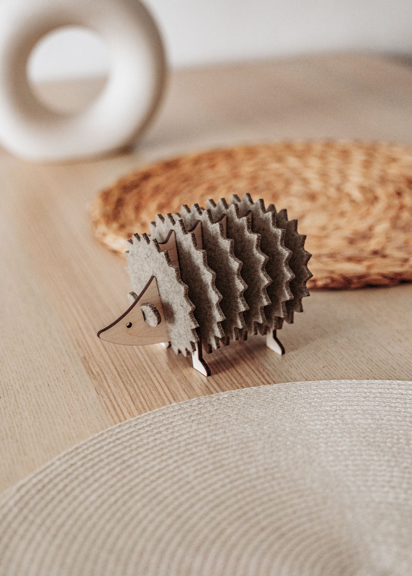 A small, decorative wooden hedgehog coaster holder sits on a wooden table with a woven placemat and a round, white ceramic object in the background, adding charm to your home decor.