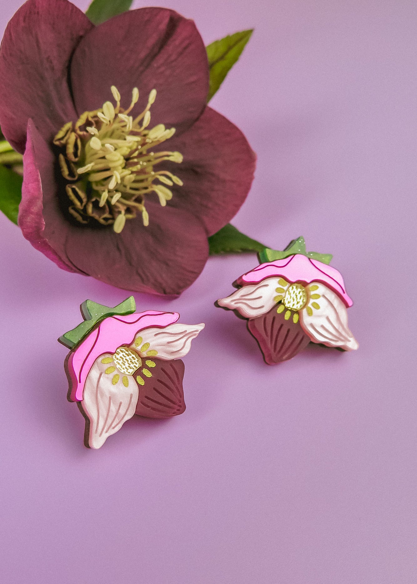 A pair of Hellebore Stud Earrings with pink, white, and green accents are displayed on a purple surface next to a real deep pink flower with yellow stamens and green leaves—perfect handcrafted floral jewelry for unique accessories lovers.