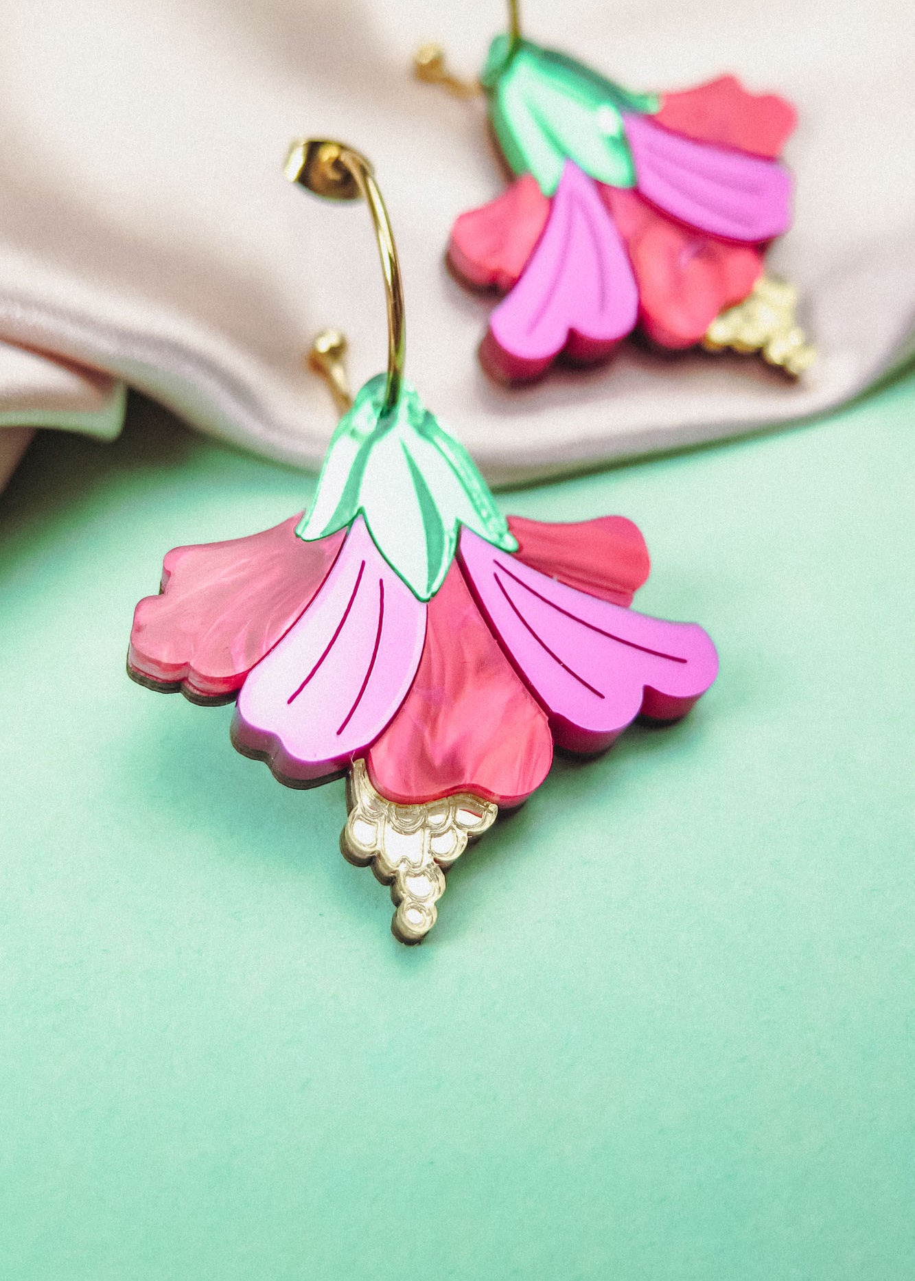 A close-up of handcrafted Hibiscus Earrings, featuring a tropical statement flower with pink and magenta petals, green leaves, and gold-tone details, resting on a mint green surface near soft beige fabric.