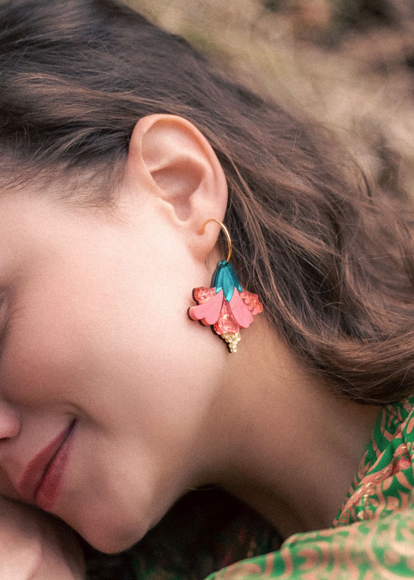 A woman with wavy brown hair wears vibrant Hibiscus Earrings—gold hoops adorned with pink, teal, and gold floral charms. She is smiling softly, her tropical statement green patterned clothing visible.