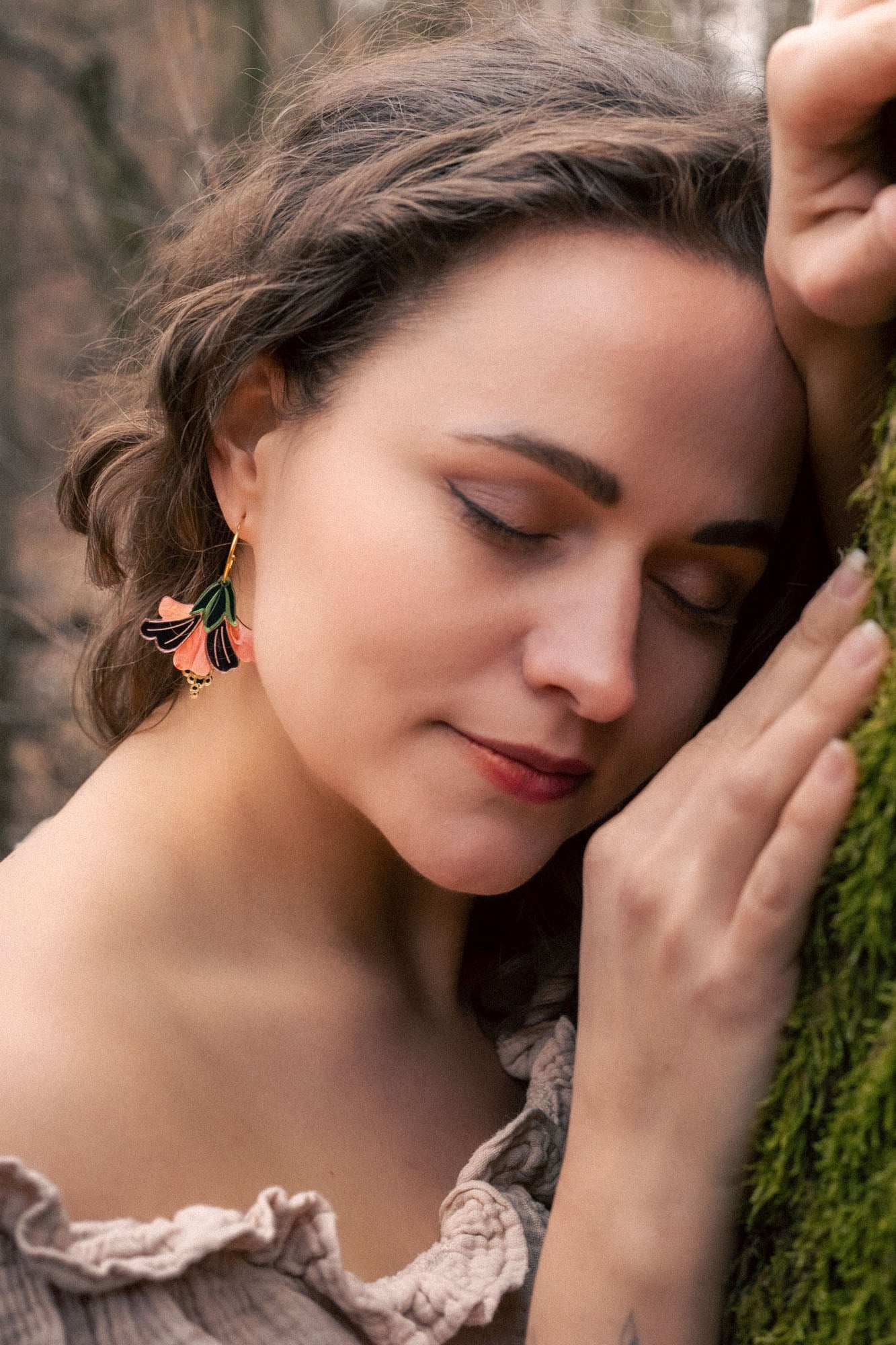 A woman with wavy brown hair and handcrafted Hibiscus Earrings gently leans her head and hand against a mossy tree, eyes closed, wearing an off-shoulder blouse—radiating a tropical statement in a serene forest setting.