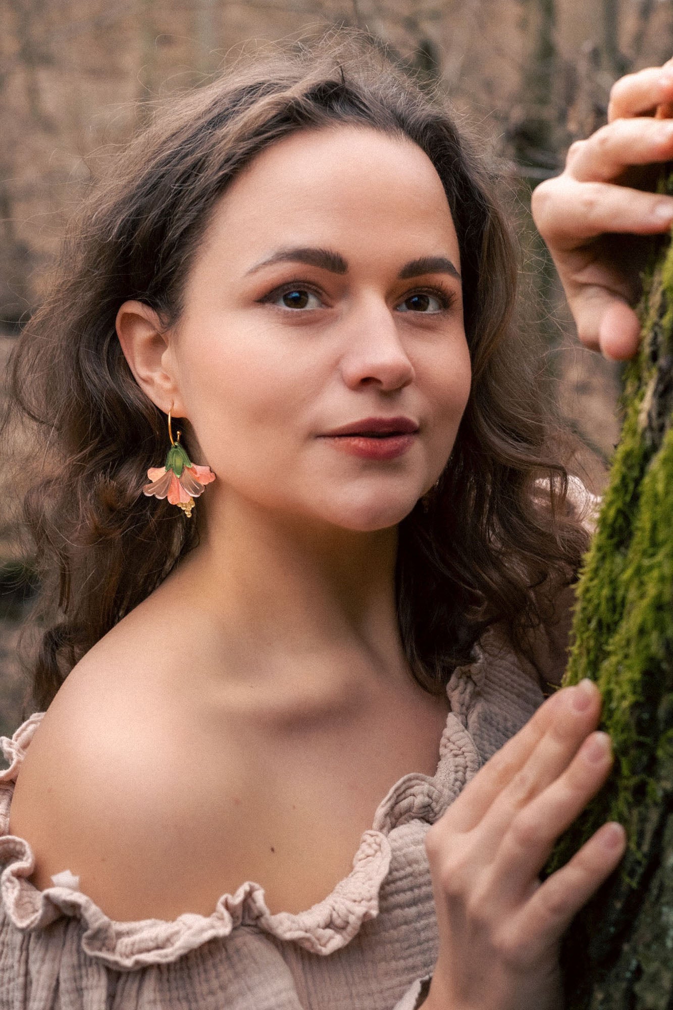 A woman with wavy brown hair and floral handcrafted Hibiscus Earrings stands in a forest, wearing an off-shoulder beige top. She gently holds a mossy tree trunk and looks at the camera with a soft, calm expression.