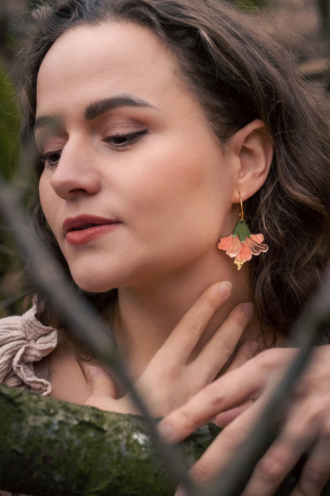 A woman with wavy brown hair and handcrafted Hibiscus Earrings gently touches her neck while standing in a forest. Her eyes are closed, branches cross before her, and she wears a beige top with ruffled details.