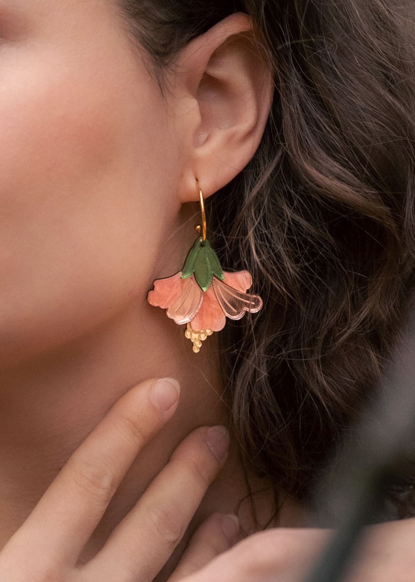 Close-up of a woman with wavy brown hair wearing handcrafted Hibiscus Earrings shaped like a pink and green flower, touching her neck gently with her fingers for a tropical statement look.