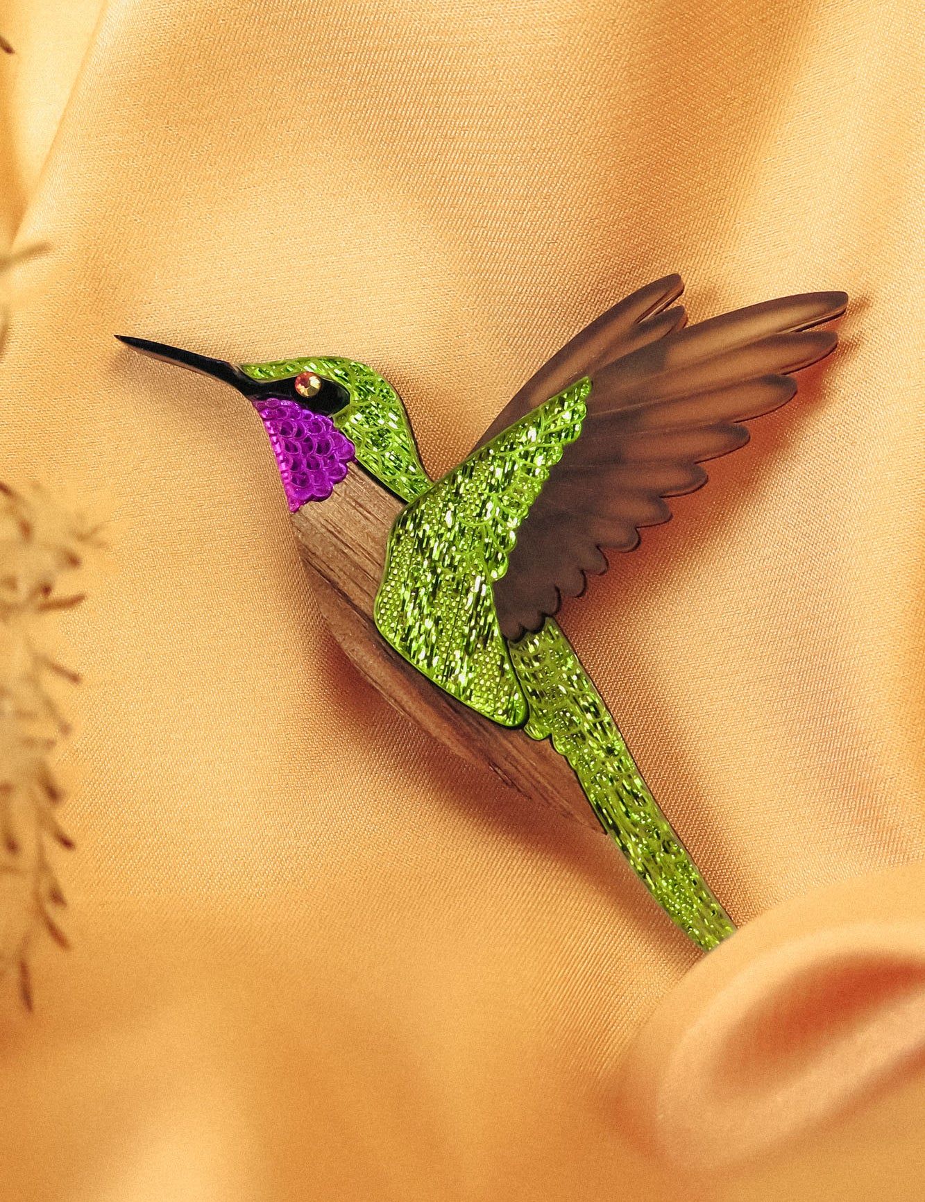 A handcrafted hummingbird brooch with vibrant green and purple glittery hues and brown wings rests on soft, peach-colored fabric.
