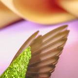 Close-up of a handcrafted green bird wing with intricate texture, featuring vibrant colors, set against a softly blurred pastel background with shades of pink, cream, and purple—perfect for a unique Hummingbird Brooch.