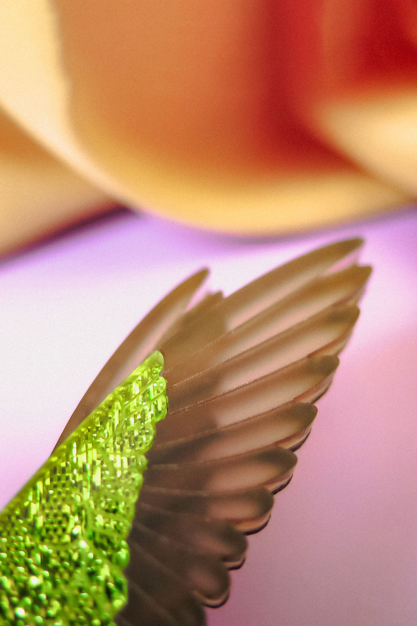 Close-up of a handcrafted green bird wing with intricate texture, featuring vibrant colors, set against a softly blurred pastel background with shades of pink, cream, and purple—perfect for a unique Hummingbird Brooch.
