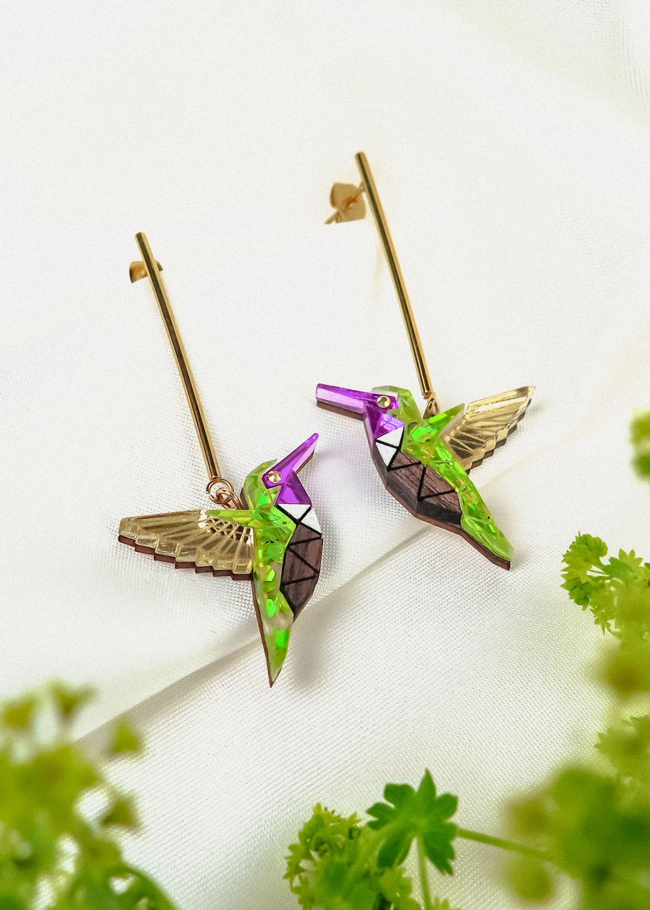 A pair of colorful, handcrafted Hummingbird Earrings with geometric patterns and gold wings, displayed on a white fabric background with green foliage in the foreground.