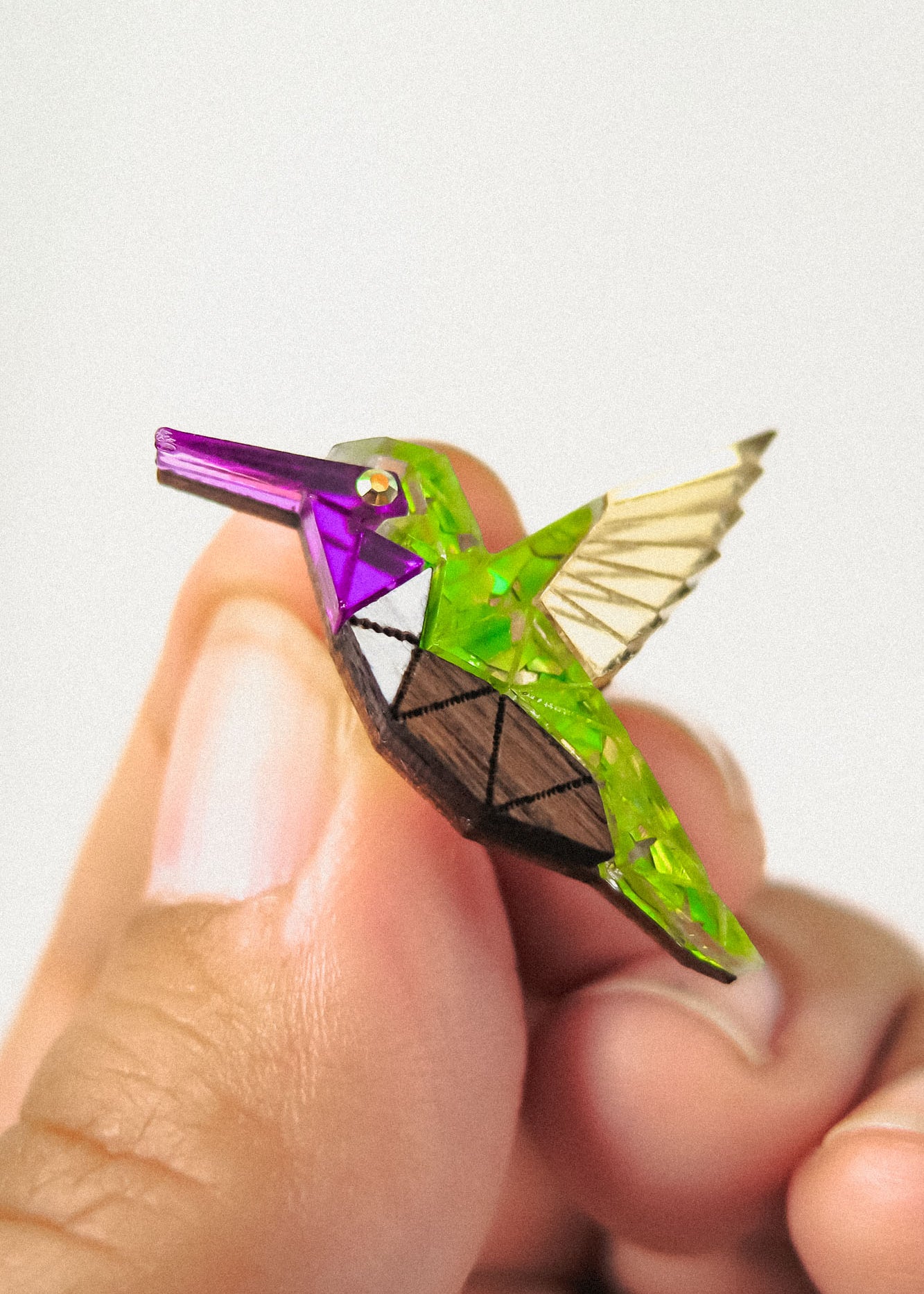 A hand holds a small, colorful hummingbird pin featuring a geometric, nature-inspired design with green, purple, gold, and brown sections and transparent wings.