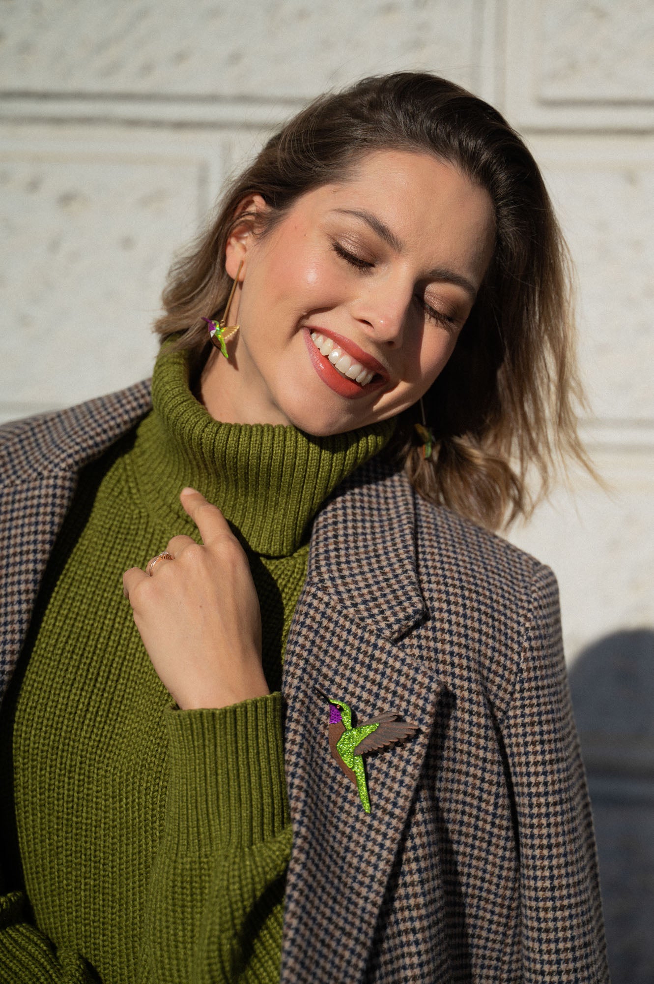 A smiling woman in a green turtleneck and plaid jacket stands against a light wall, wearing handcrafted Hummingbird Earrings and matching lapel jewelry. Sunlight highlights her face as she closes her eyes, radiating joy and sustainable craftsmanship.