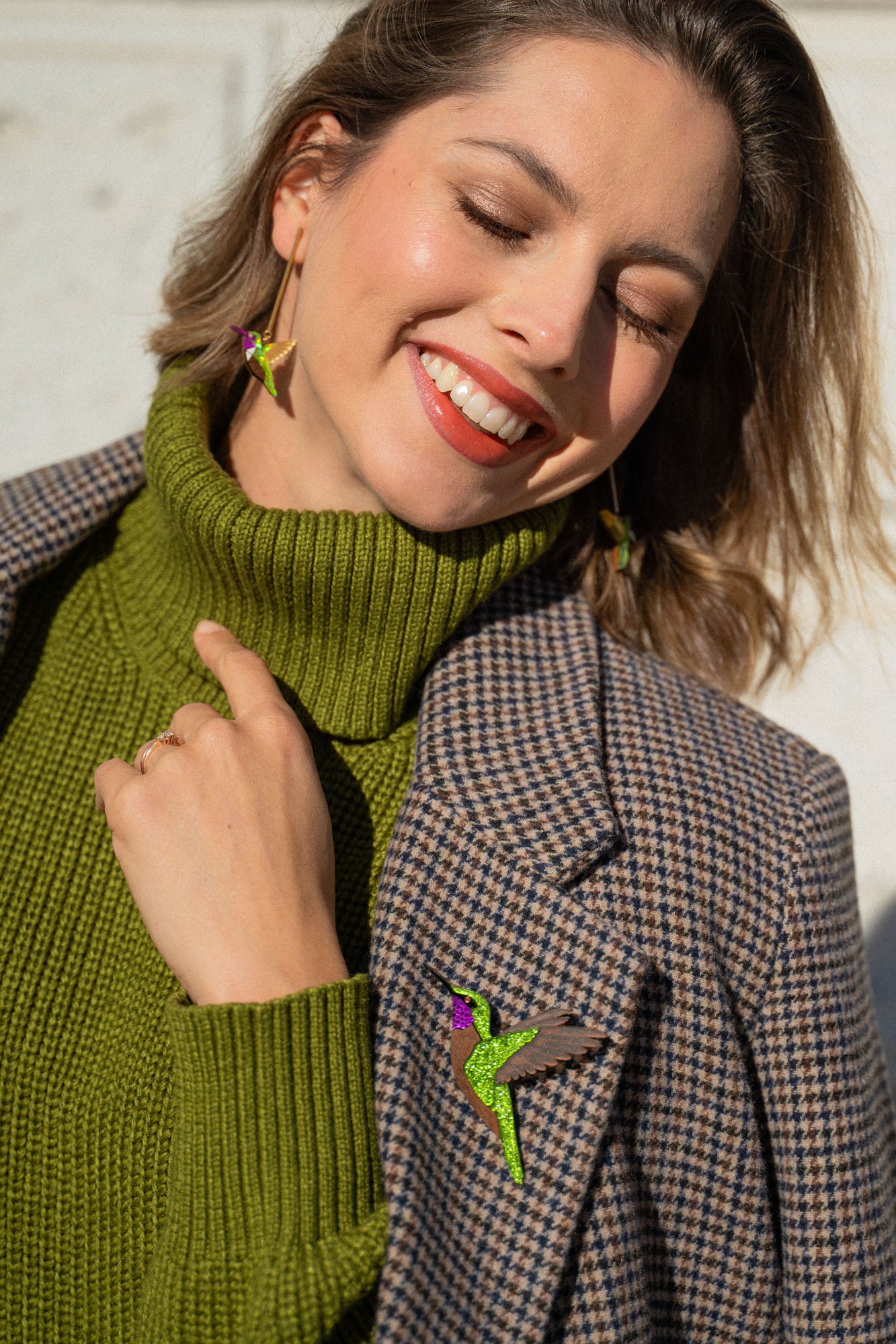 A smiling woman in a green turtleneck and plaid blazer stands in sunlight, eyes closed and head tilted. She wears bird-shaped earrings and a handcrafted Hummingbird Brooch, all made from premium materials.