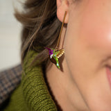 Close-up of a woman's ear wearing a long gold Hummingbird Earring, handcrafted with green, gold, and purple details. She pairs it with a green knitted turtleneck sweater, showcasing sustainable craftsmanship.