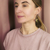A woman with light brown hair in a ponytail wears a light pink shirt, a colorful hummingbird earring, and a nature-inspired hummingbird pin while standing in front of a pinkish curtain.