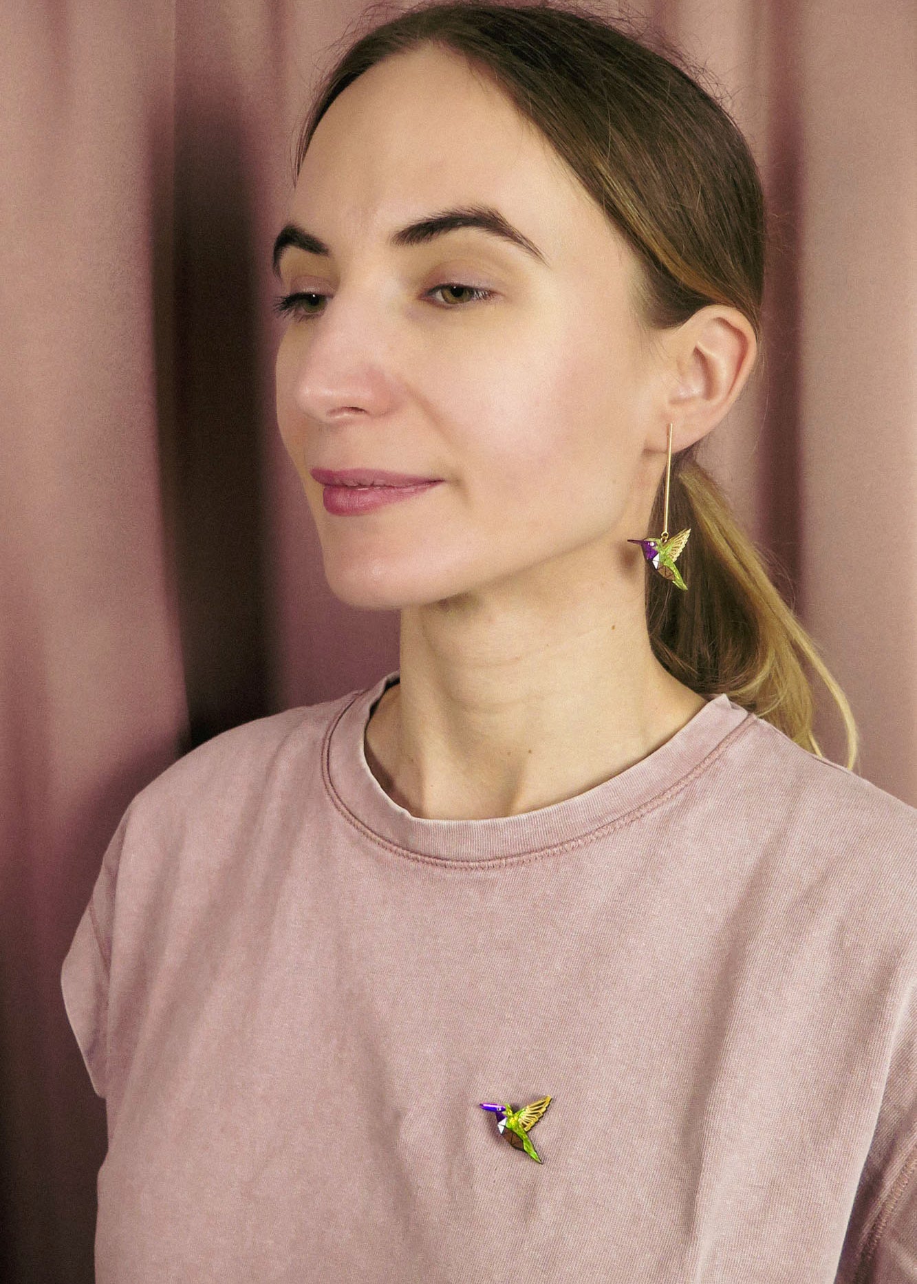 A woman with light brown hair in a ponytail wears a light pink shirt, a colorful hummingbird earring, and a nature-inspired hummingbird pin while standing in front of a pinkish curtain.