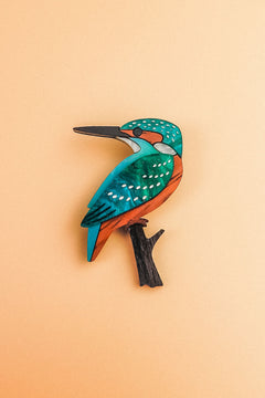 A colorful, stylized Kingfisher Brooch handcrafted from sustainable materials, with teal, orange, and white markings perched on a small black branch, set against a pale peach background.
