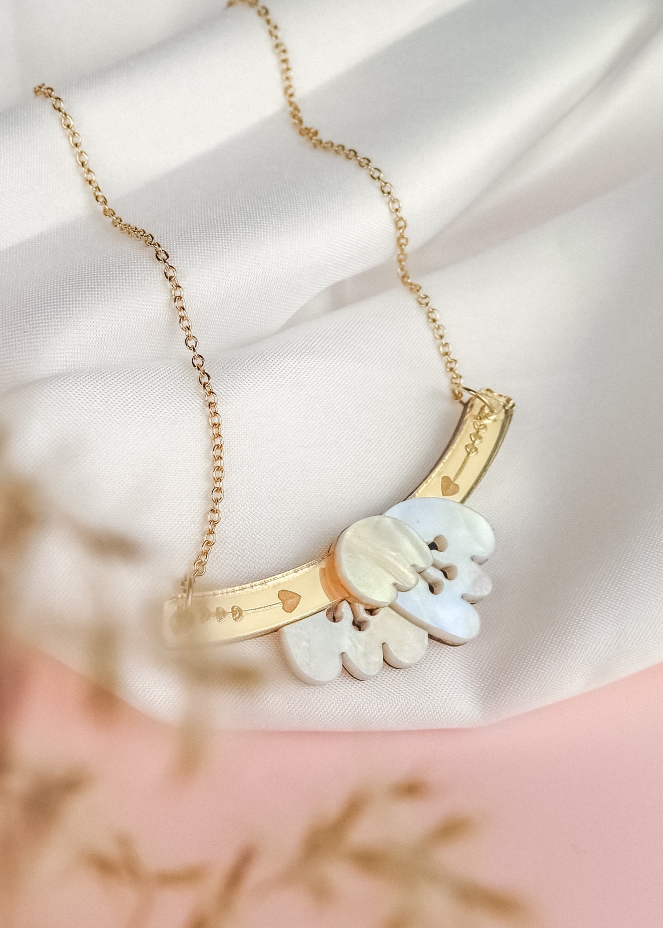 A gold Tulip Necklace with a delicate chain, featuring an intricate pendant with white and gold elements and small heart motifs, exudes romantic charm as it rests on a soft white fabric background.